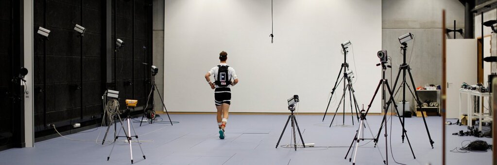 Image of someone running in the Ghent University Sport Labratory. He is wearing a sensor on the shin and a trail backpack.