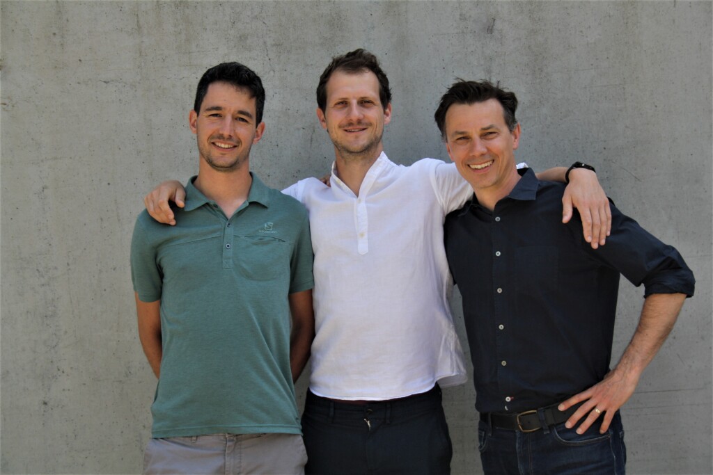 Group picture of a smiling Rud, Senne and Kristof in front of a grey wall.