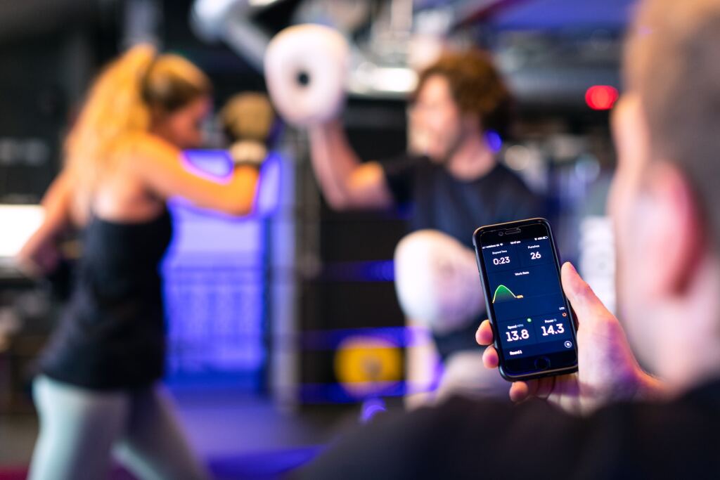 A man is holding a smartphone that shows graphs and variables. In the background are a woman and a man boxing.